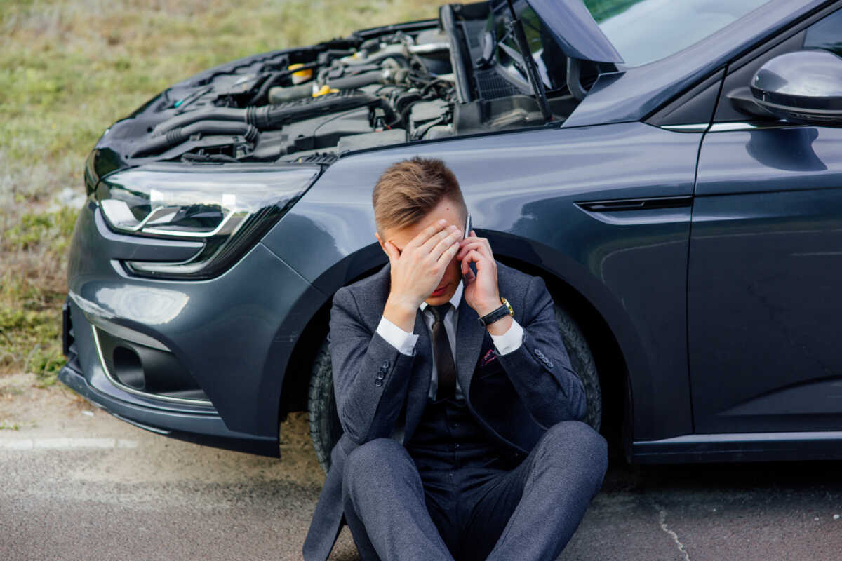 Upset young man use phone sitting on road near the broken car opened the hood help repair stress problem emergency insurance auto