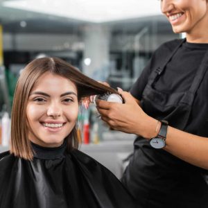 salon services, hairdresser with round brush styling hair of female customer, happy brunette woman